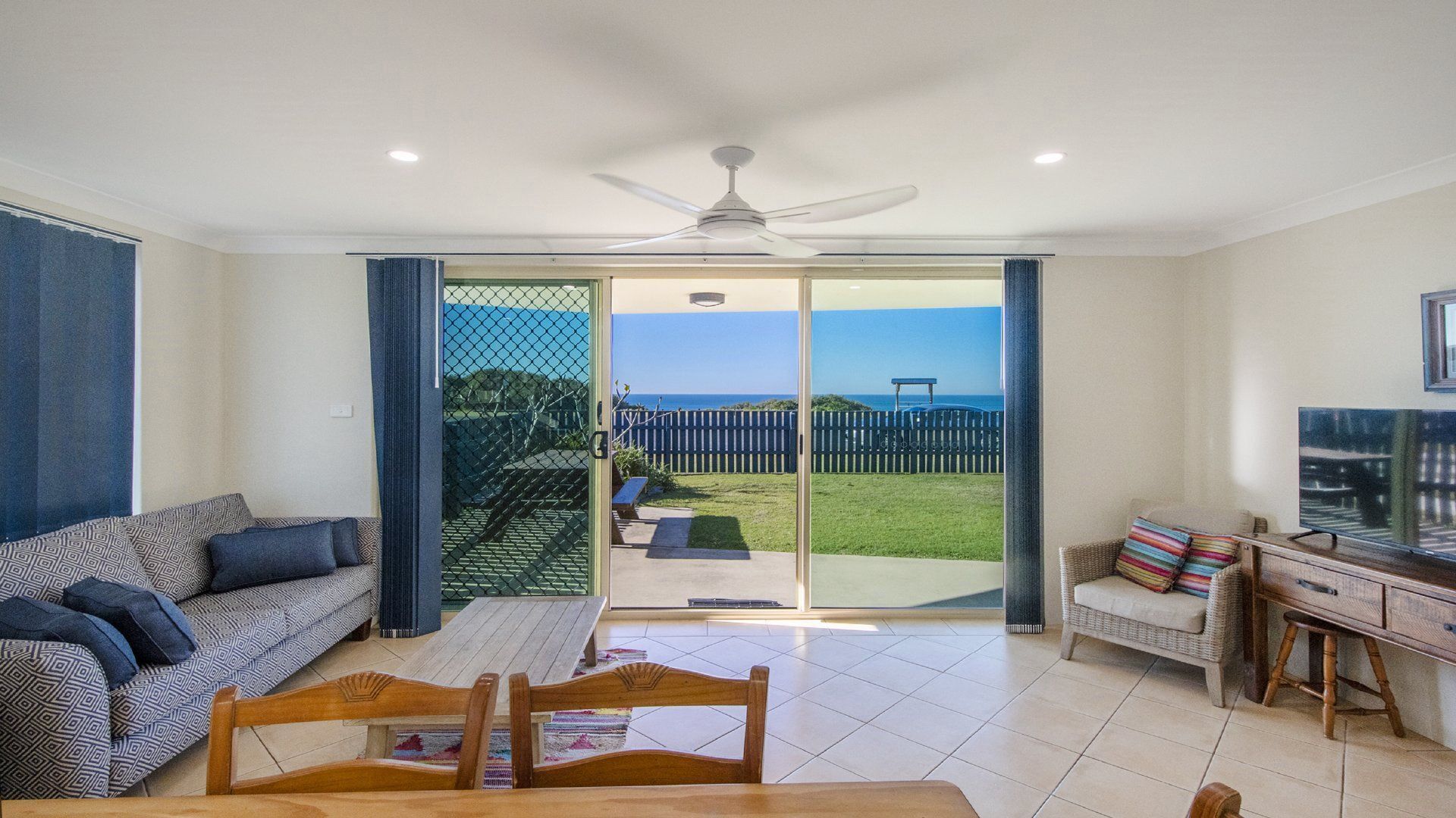 A living room with a sofa, a dining table, and a TV. There is a sliding glass door leading to a balcony with a view of the ocean.