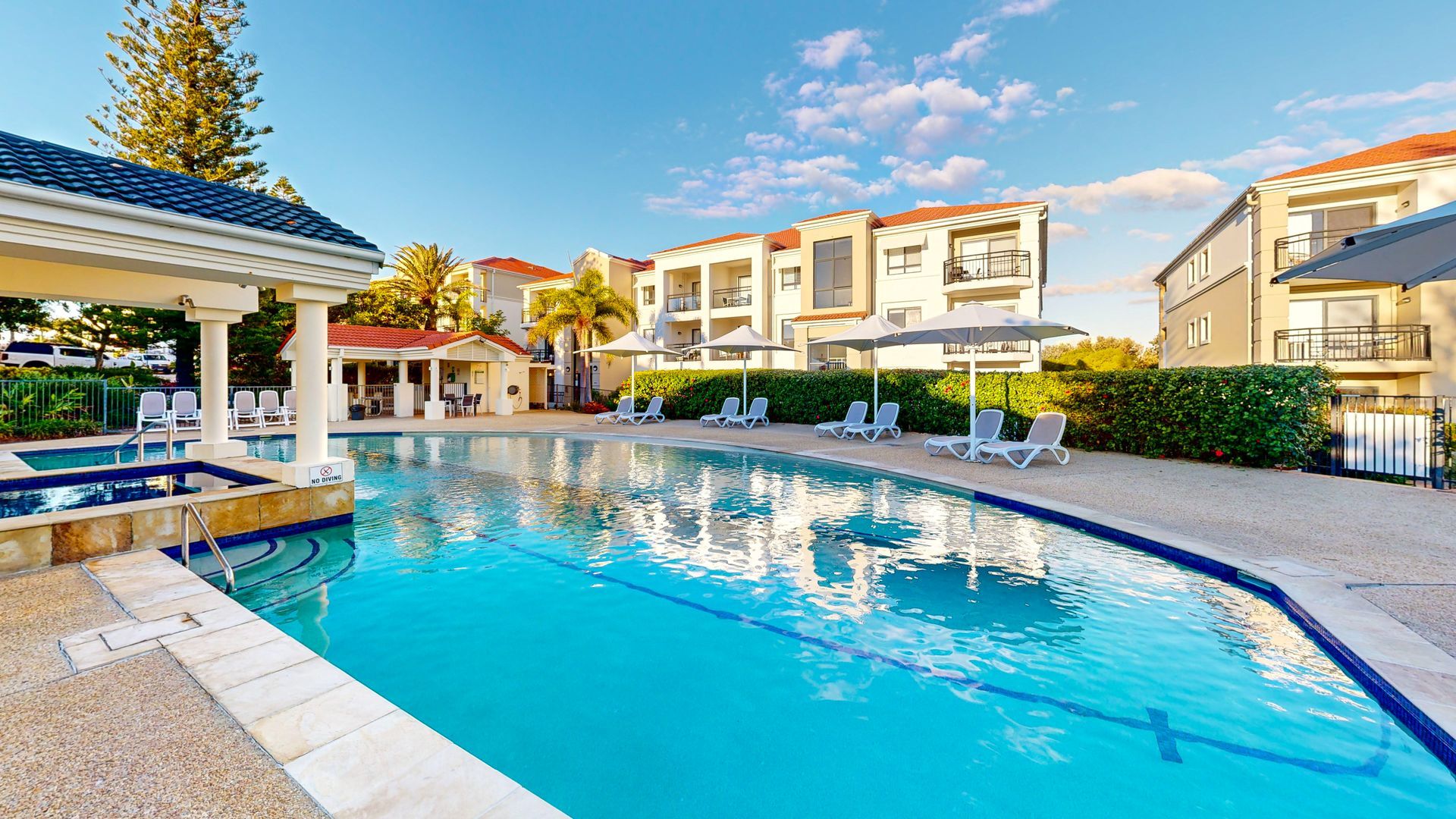 A swimming pool with lounge chairs and umbrellas, surrounded by buildings with balconies.