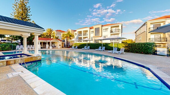 A swimming pool with lounge chairs and umbrellas, surrounded by buildings with balconies.