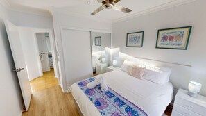 Un dormitorio con cama, mesitas de noche, ventilador de techo y dos cuadros en la pared.