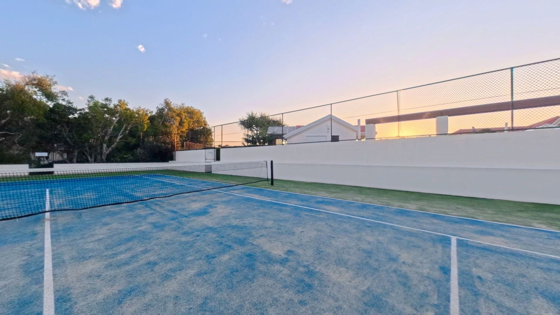 An outdoor tennis court with a net, surrounded by a white wall and trees.