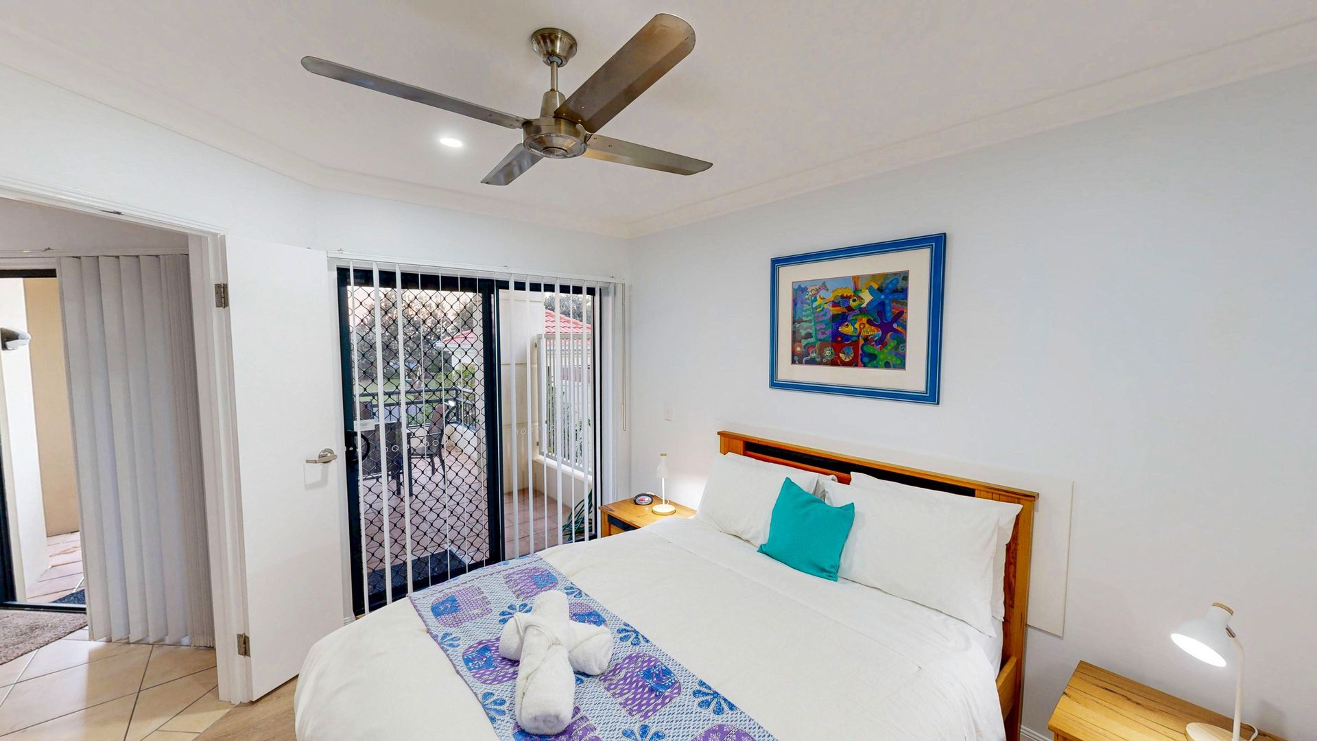 A bedroom with a bed, a ceiling fan, a nightstand, a lamp, a painting, and a view through a door to an outdoor area.