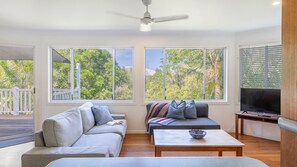 A living room with a sofa, a coffee table, and a TV. There are large windows with a view of trees and a balcony.