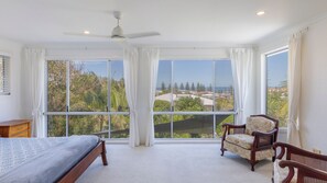 A bedroom with a bed, a chair, a ceiling fan, and large windows offering a view of the outdoors.