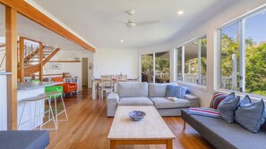 A spacious living area with a wooden staircase, a dining area, and large windows offering a view of greenery.