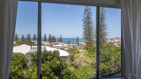 A view through a window with curtains, overlooking a coastal area with houses, trees, and the sea.