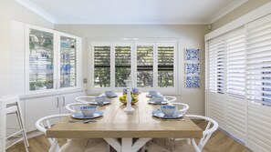 A dining area with a wooden table, white chairs, and a window with blinds. - Marina Views - Lifestyle Yamba (YAMBA)