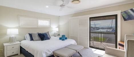 A bedroom with a bed, bedside tables, a bench, a chair, a ceiling fan, and a view of a marina through a sliding door.