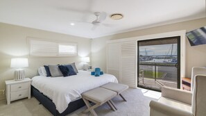 A bedroom with a bed, bedside tables, a bench, a chair, a ceiling fan, and a view of a marina through a sliding door.