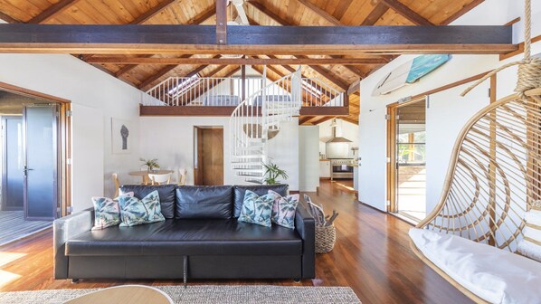 A spacious living area with a black leather sofa, a wooden ceiling, and a staircase leading to an upper level.