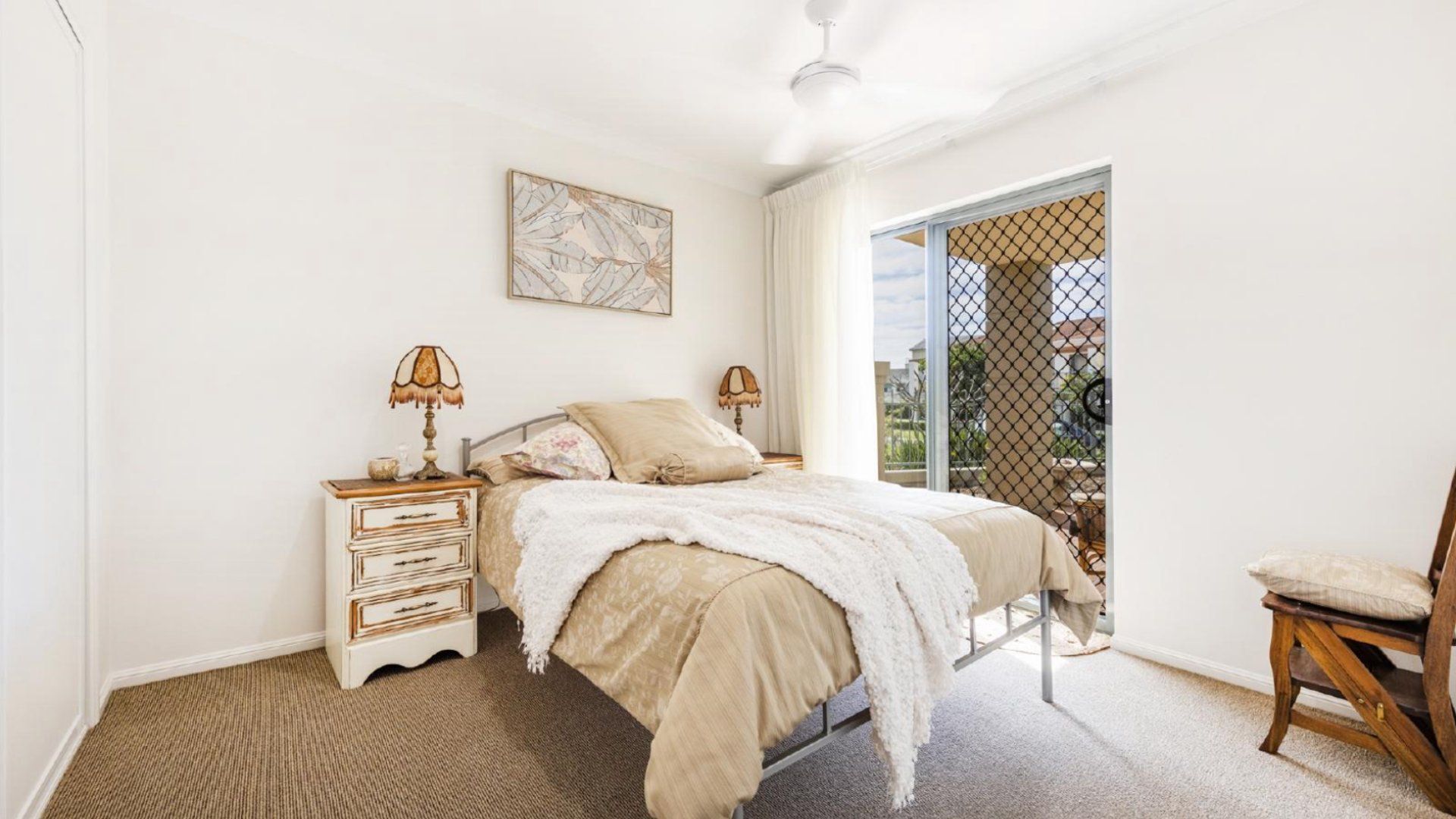 A bedroom with a bed, two bedside tables with lamps, a chair, a window with a view, and a ceiling fan.
