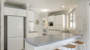 A modern kitchen with white cabinetry, a built-in microwave, and a large island with bar stools. - Yamba Family Seascape - Lifestyle Yamba (YAMBA)