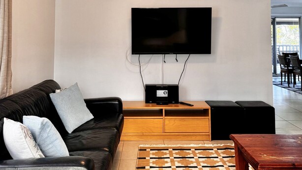 A living room with a black leather sofa, a flat-screen TV mounted on the wall, a wooden coffee table, and a patterned rug. - The Perch - Lifestyle Yamba (YAMBA)