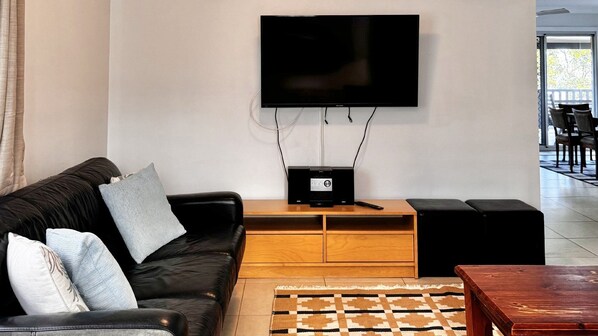A living room with a black leather sofa, a flat-screen TV mounted on the wall, a wooden coffee table, and a patterned rug.