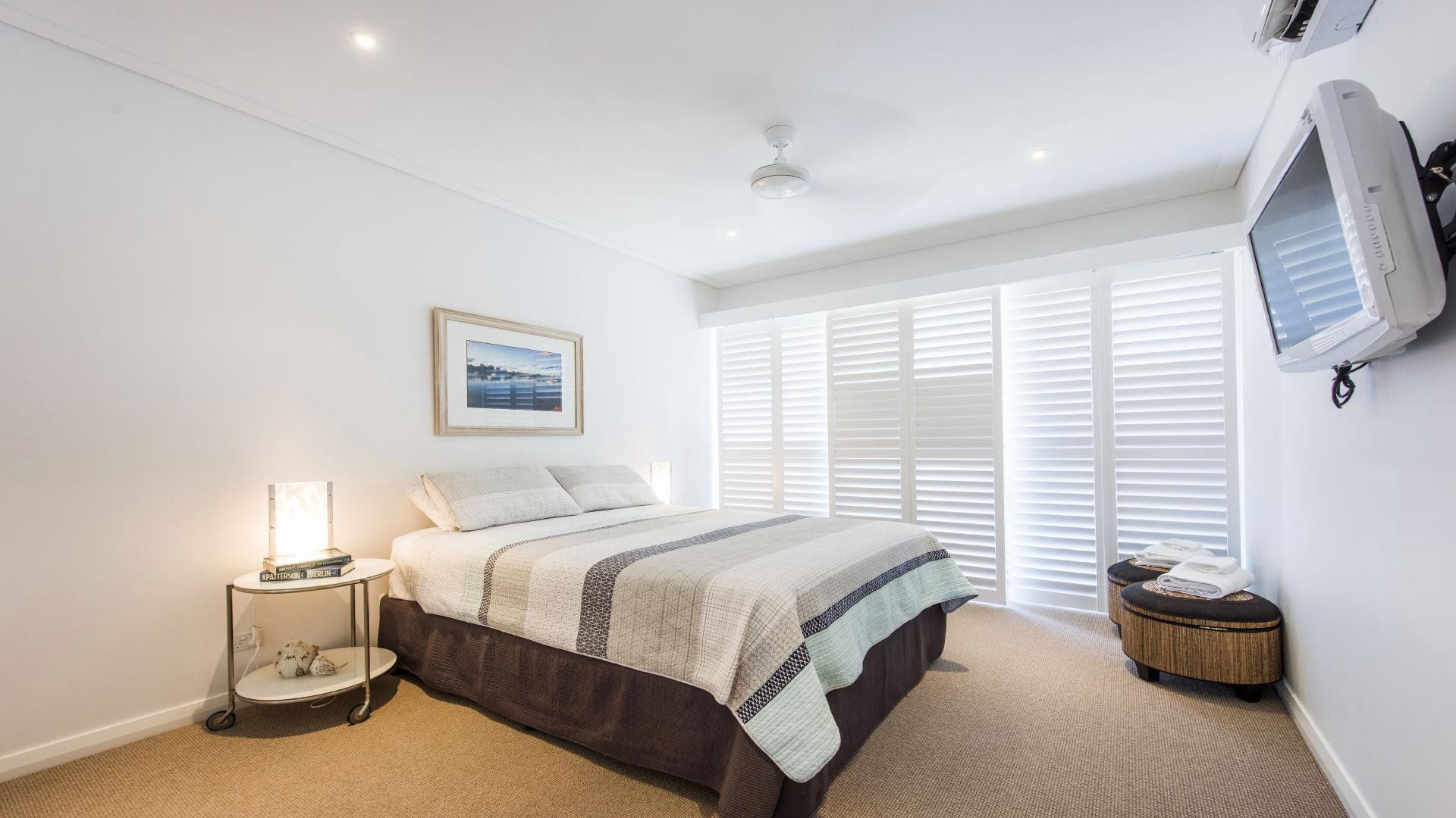 A bedroom with a bed, a bedside table with a lamp, a picture on the wall, a window with blinds, and a television mounted on the wall.