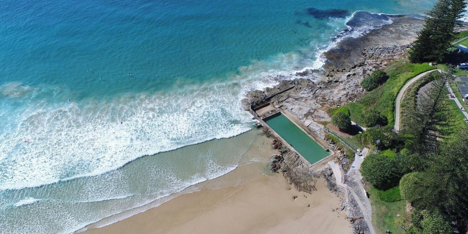 An aerial view of a coastal area with a swimming pool, beach, and a walking path.