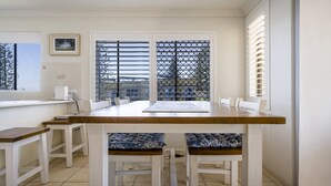 Comedor con mesa de madera y sillas blancas, una ventana con persianas y vistas al exterior.