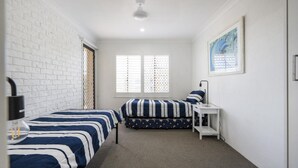 Una habitación con dos camas, una ventana con persianas, una mesita y un ventilador de techo.