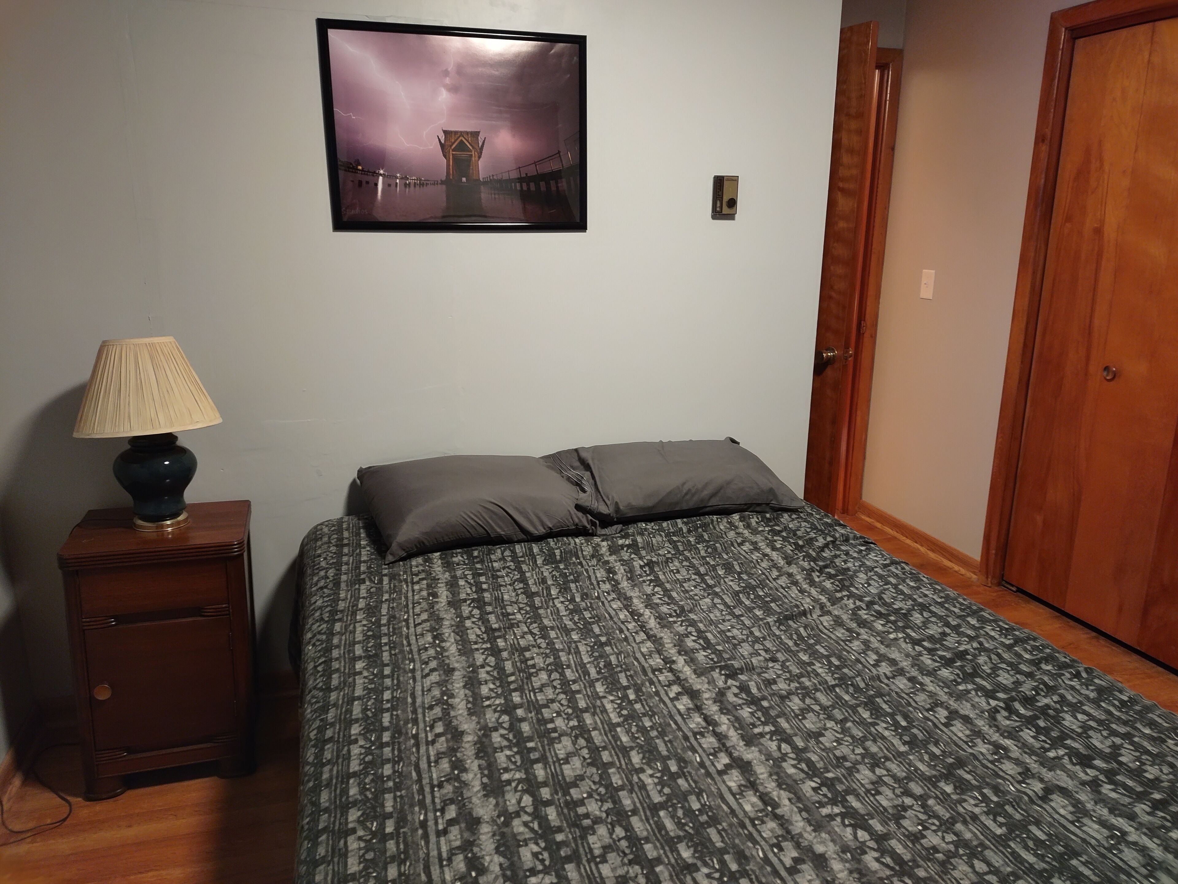 A bedroom with a bed, a nightstand with a lamp, a framed picture on the wall, and a wooden door.