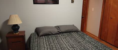 A bedroom with a bed, a nightstand with a lamp, a framed picture on the wall, and a wooden door.