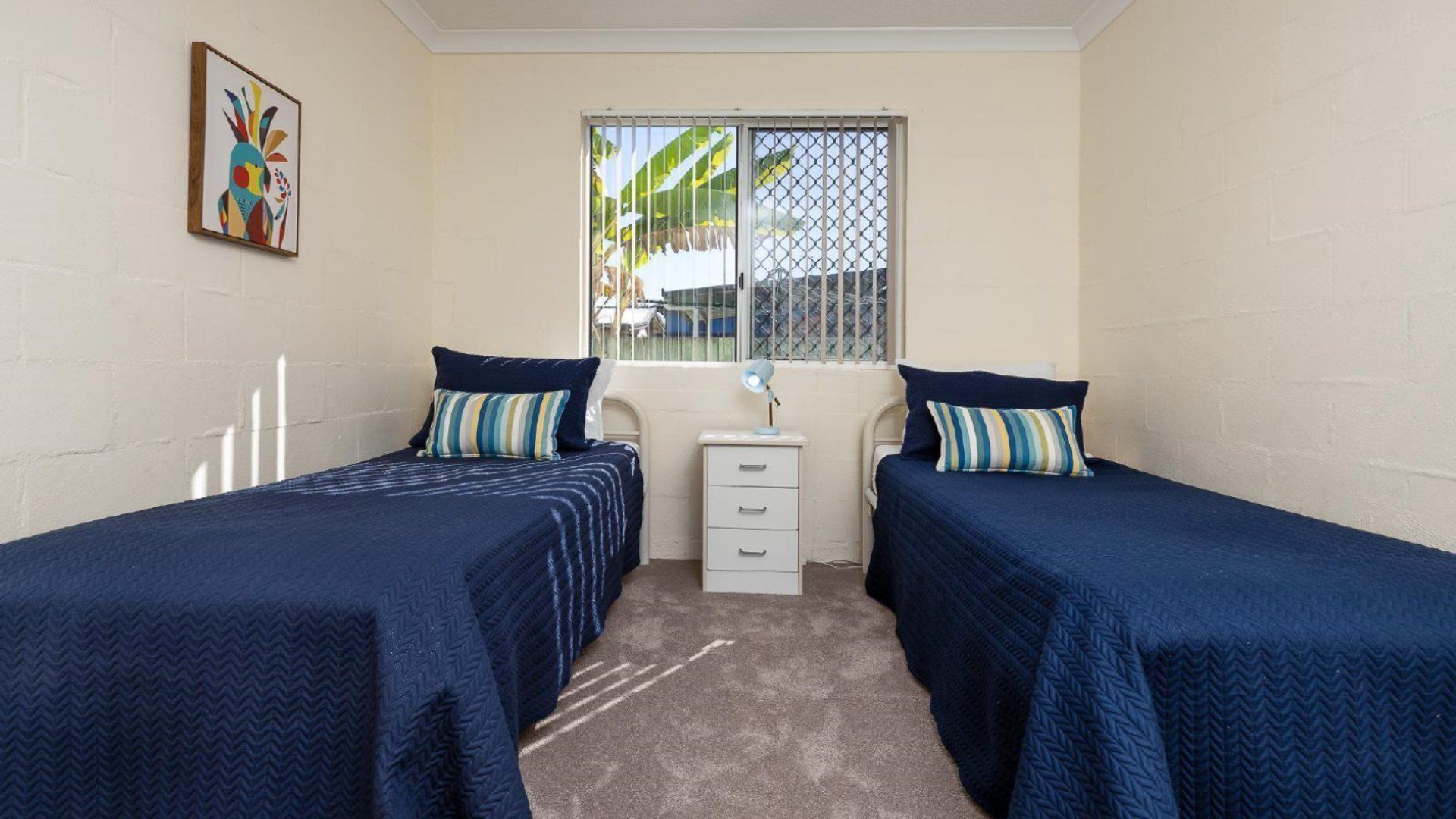 A room with two single beds, a small bedside table with a lamp, a window with a view of the outside, and a colorful painting on the wall.