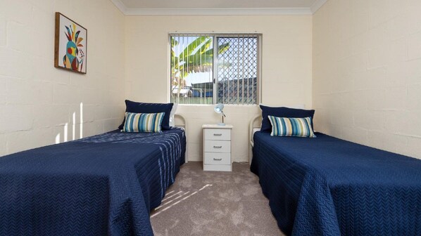 A room with two single beds, a small bedside table with a lamp, a window with a view of the outside, and a colorful painting on the wall.