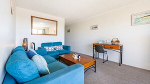 A living room with a blue sofa, a wooden coffee table, a desk with a chair, and a mirror on the wall. - Yamba Towers Unit 12 - Lifestyle Yamba (YAMBA)