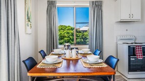 A dining area with a wooden table set for six, a window with curtains, and a kitchen with a stove and oven. - Yamba Towers Unit 12 - Lifestyle Yamba (YAMBA)