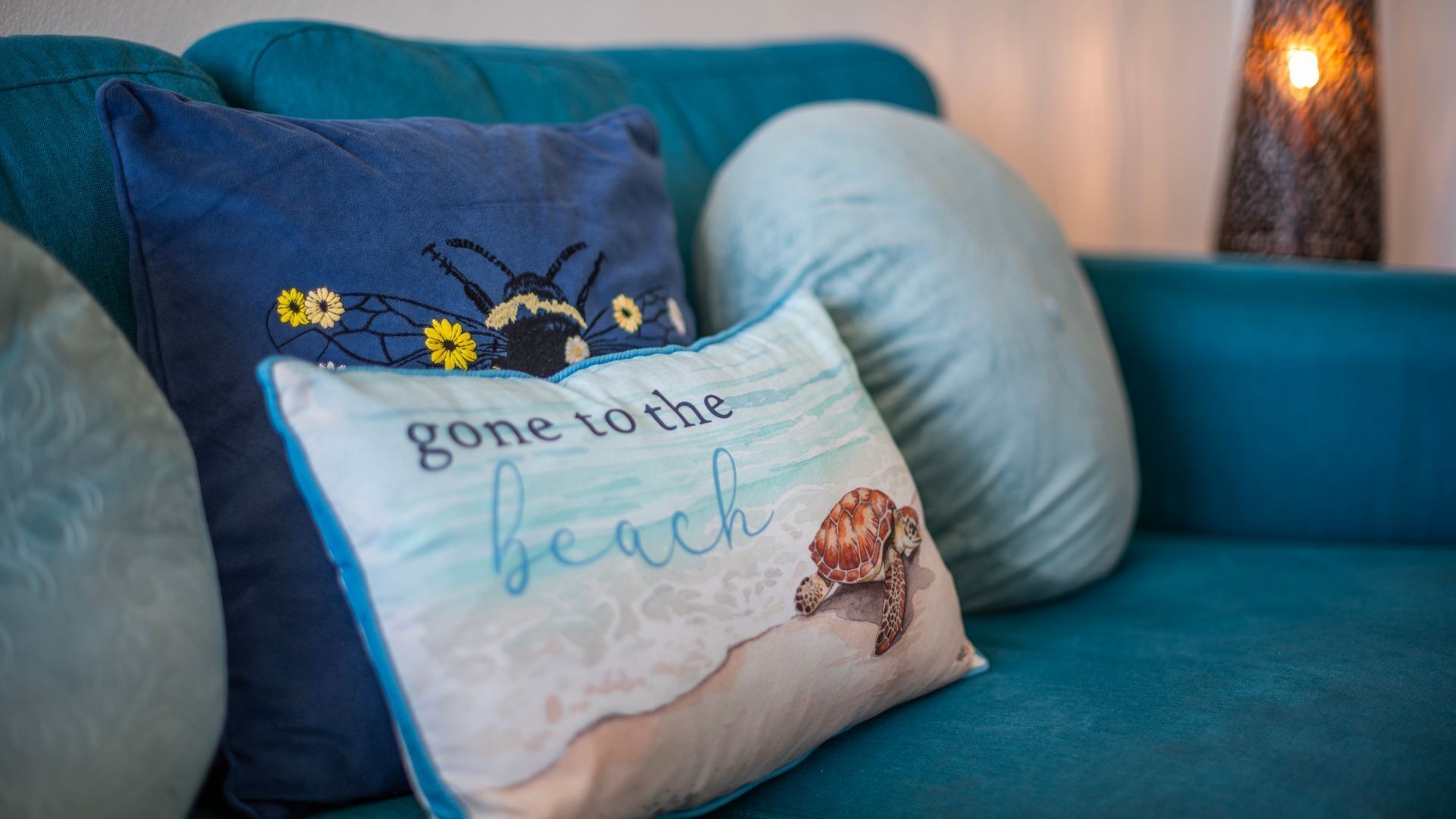 A teal sofa with decorative pillows, one featuring a beach theme with a turtle illustration and the phrase 'Gone to the beach.'