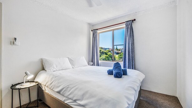 A hotel room with a bed, a bedside table with a lamp, and a window with curtains. - Yamba Towers Unit 12 - Lifestyle Yamba (YAMBA)