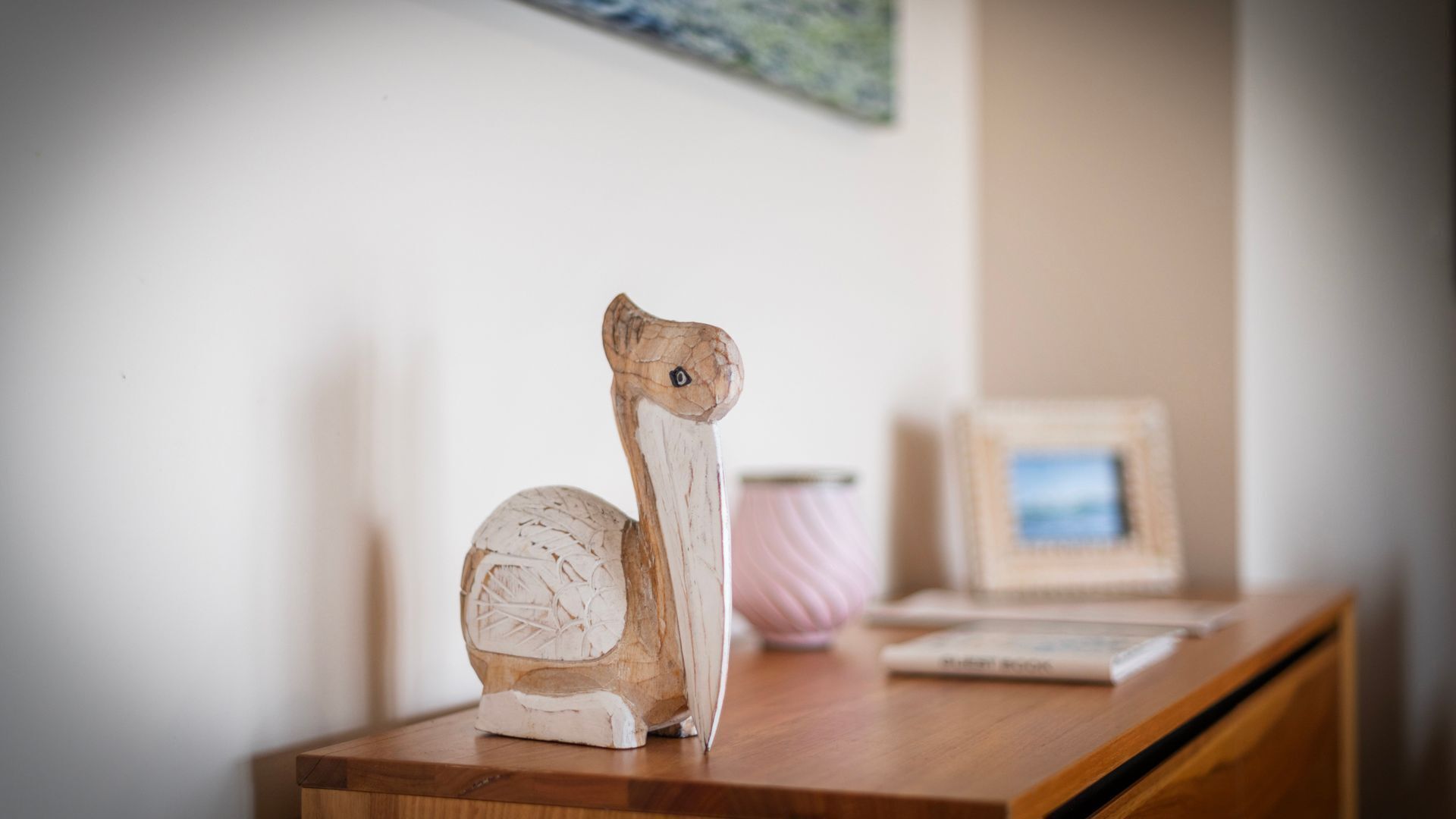 A wooden sculpture on a wooden table with a framed picture and a pink vase in the background.