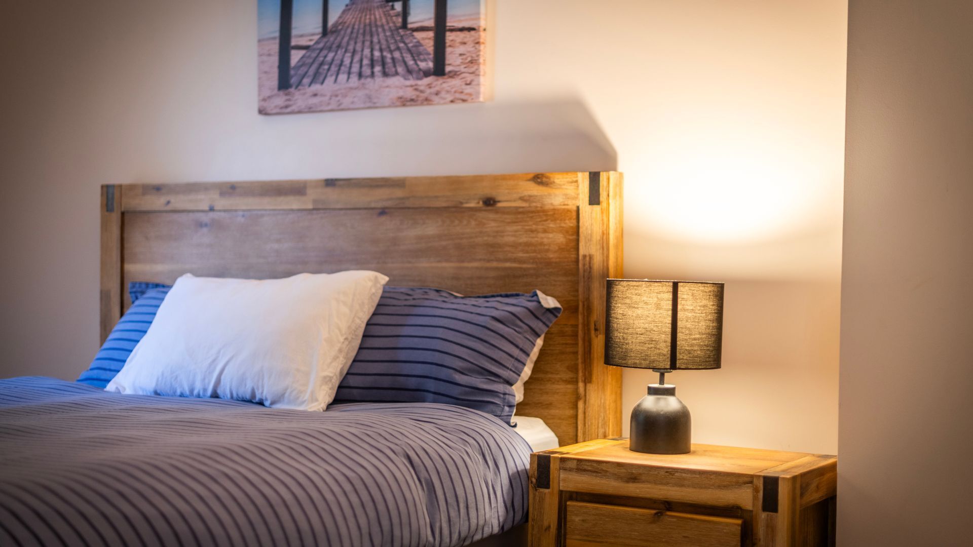 A bed with a wooden headboard, a striped bedspread, and a pillow. There is a nightstand with a lamp beside the bed.