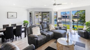 A modern living room with a grey sectional sofa, a dining area with a table and chairs, and a large window offering a view of the outdoors. - Anchorage Waterfront Unit - Lifestyle Yamba (YAMBA)