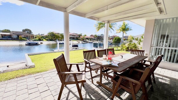 A covered patio with a table and chairs, overlooking a body of water with boats and houses in the background. - Anchorage Waterfront Unit - Lifestyle Yamba (YAMBA)