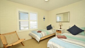 Quarto com duas camas de solteiro, uma cadeira, uma mesa de cabeceira com abajur e uma janela com persianas.