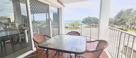 Un balcon avec une table en verre et des chaises en osier, donnant sur une zone côtière.