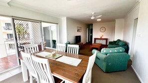 A dining area with a wooden table and white chairs, a green sofa, a television, and a ceiling fan.
