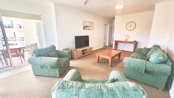 A living room with two green sofas, a wooden coffee table, a TV on a wooden stand, and a clock on the wall.
