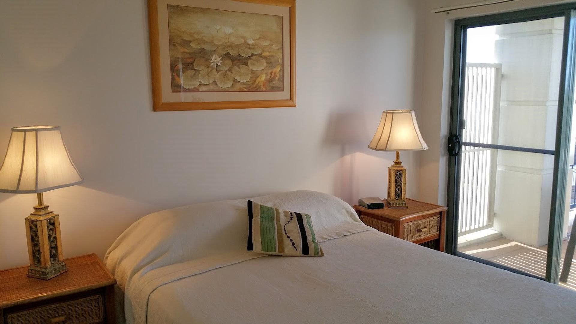 A bedroom with a bed, two bedside lamps, a pillow, a framed artwork on the wall, and a view through a glass door.