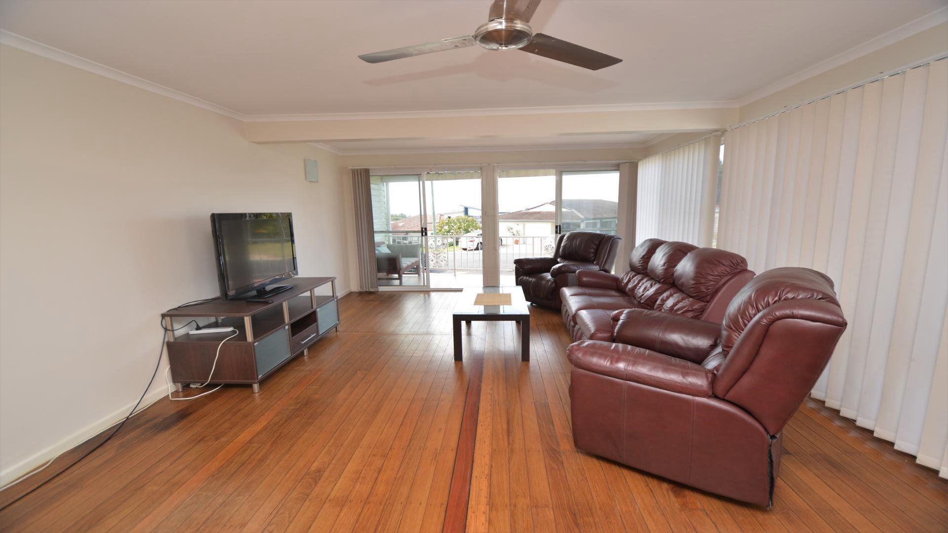A living room with a flat-screen TV, leather sofa, wooden flooring, and a ceiling fan.