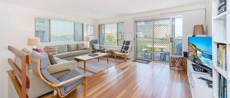 A spacious living room with a sofa, chairs, a coffee table, and a TV. There are large windows with blinds and a view of the outdoors.
