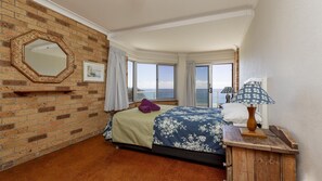 A hotel room with a brick wall, a large window with a view of the sea, a bed with a floral bedspread, and a wooden nightstand with a lamp.