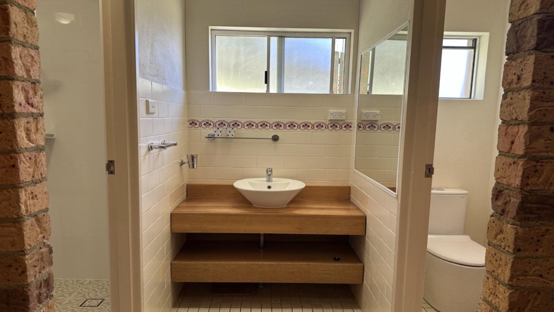 Une salle de bain avec un lavabo blanc, un meuble-lavabo en bois, des murs carrelés et une fenêtre.