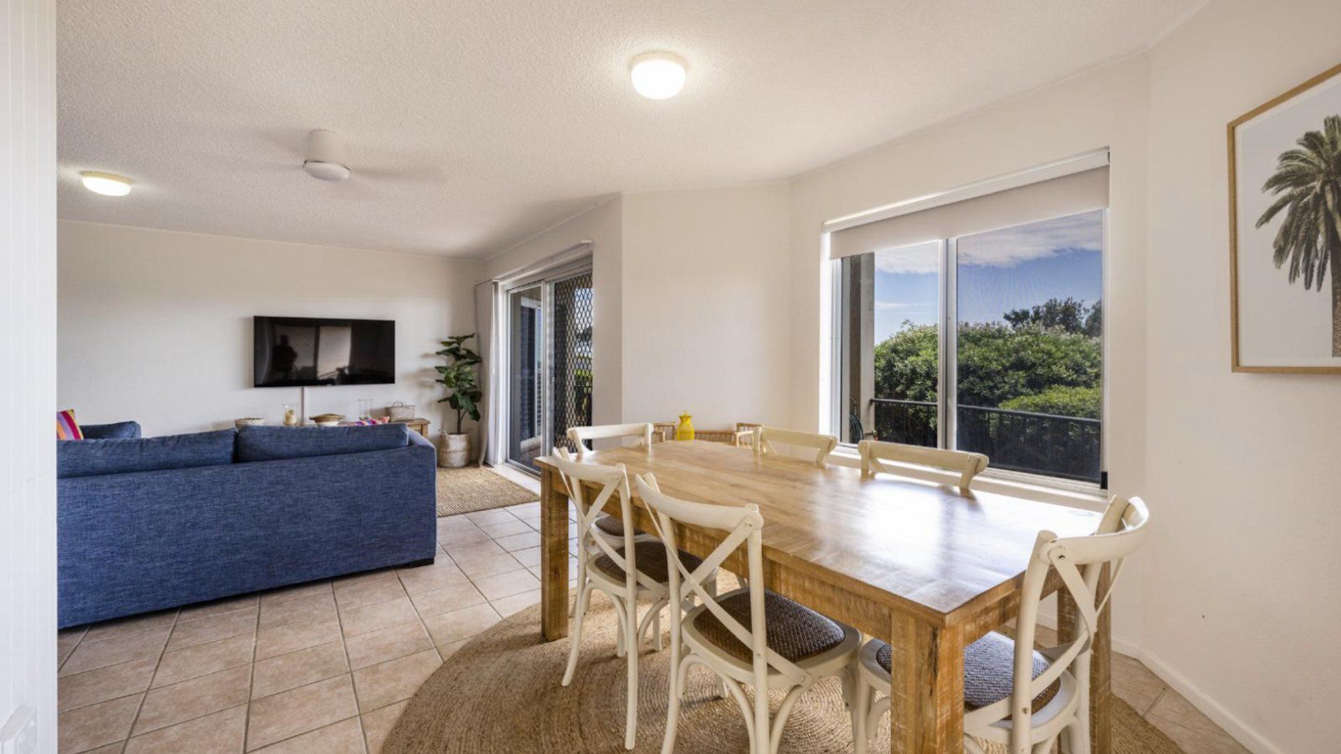 A modern living room with a blue sofa, a wooden dining table with white chairs, and a large window offering a view of greenery.