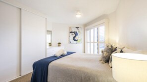 A bedroom with a bed, a bedside lamp, a window with blinds, and a wall-mounted artwork. - Romiaka Unit 1 - Lifestyle Yamba (YAMBA)