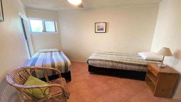Acomodação pequena, um único quarto com duas camas, uma cadeira, mesa de cabeceira e janela.