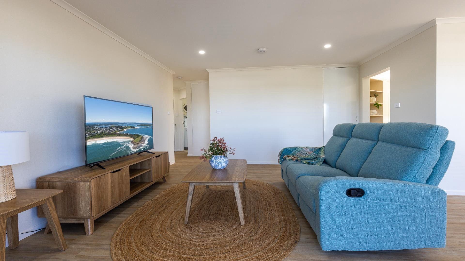 A modern living room with a flat-screen TV, a blue sofa, a wooden coffee table, and a woven rug.