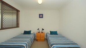 A small, clean room with two single beds, a wooden dresser, and a window with blinds.