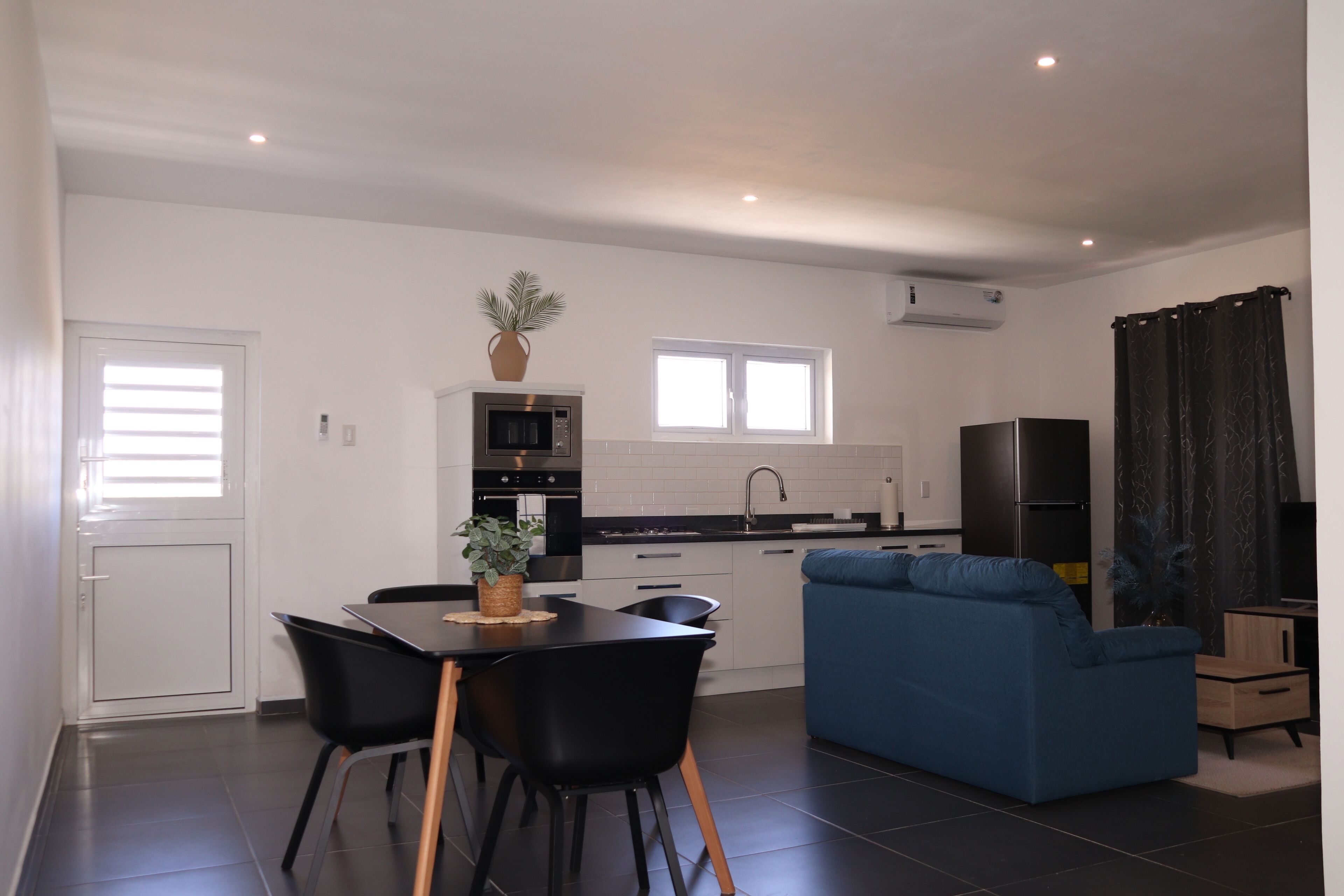 Una cocina moderna con armarios blancos, encimera negra y horno empotrado. Hay un comedor con mesa y sillas negras, y un sofá azul en la sala de estar.
