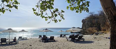 Une plage avec des chaises longues et des parasols, un ciel dégagé et une falaise rocheuse.